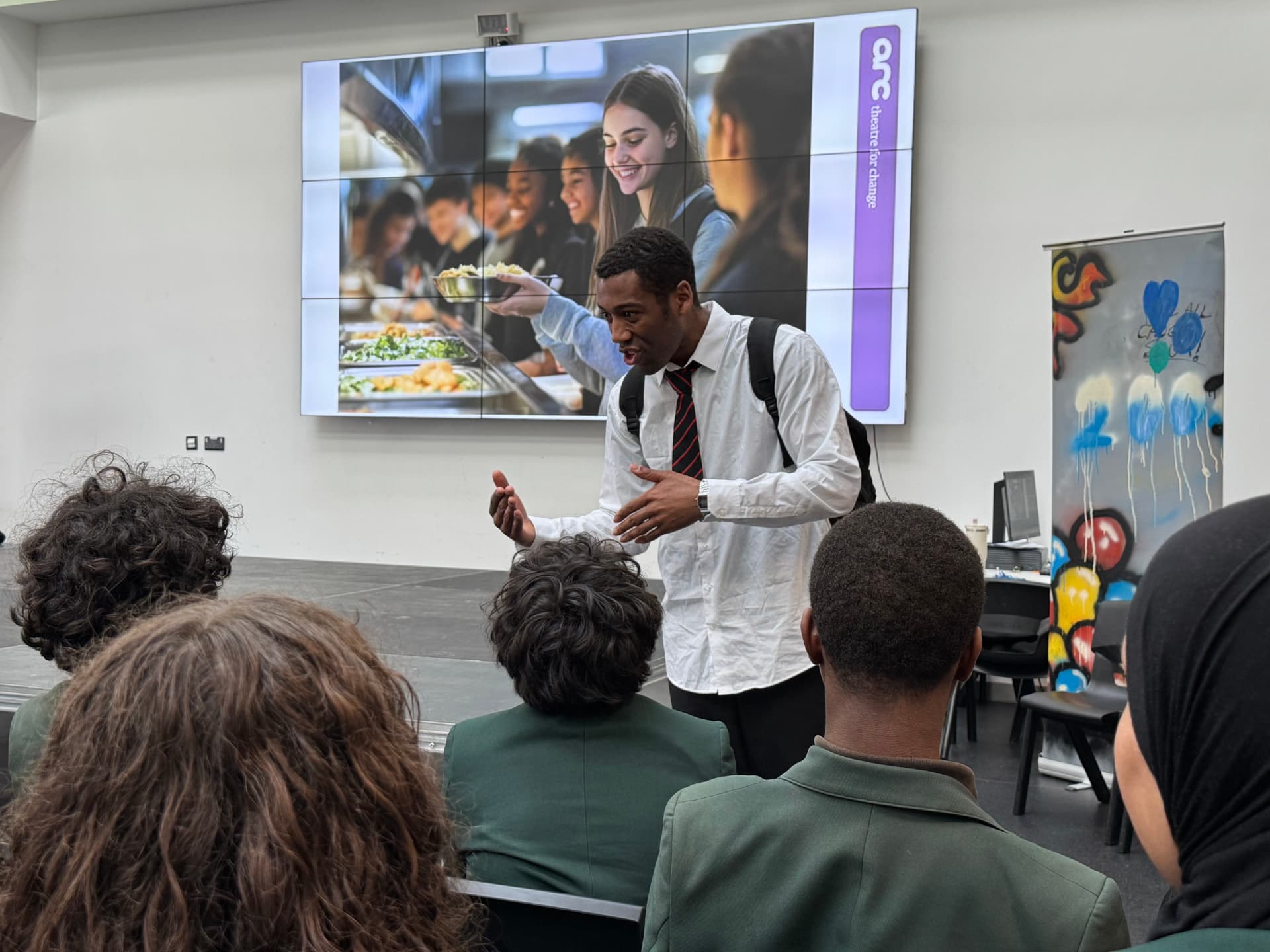Time's Up! actor addressing pupils with a school canteen image projected behind